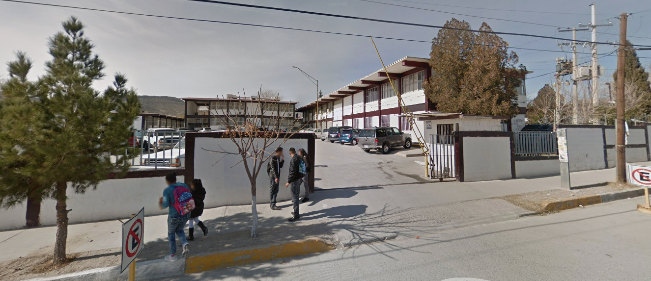 Fotografía en la que se muestra la entrada del plantel, conformada por muros blancos. En medio de los muros se encuentra el acceso al estacionamiento. Al fondo se encuentran las instalaciones.